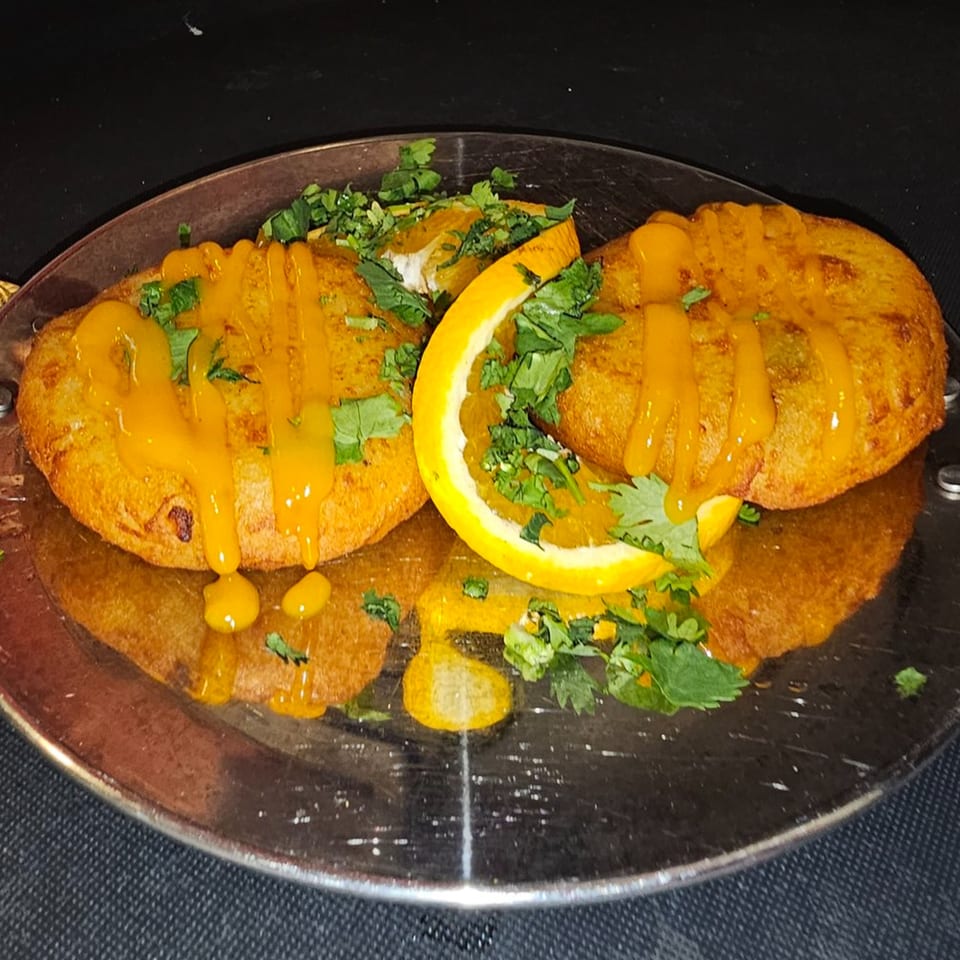 Two golden-brown Indian patties drizzled with orange sauce and garnished with cilantro and orange slices.