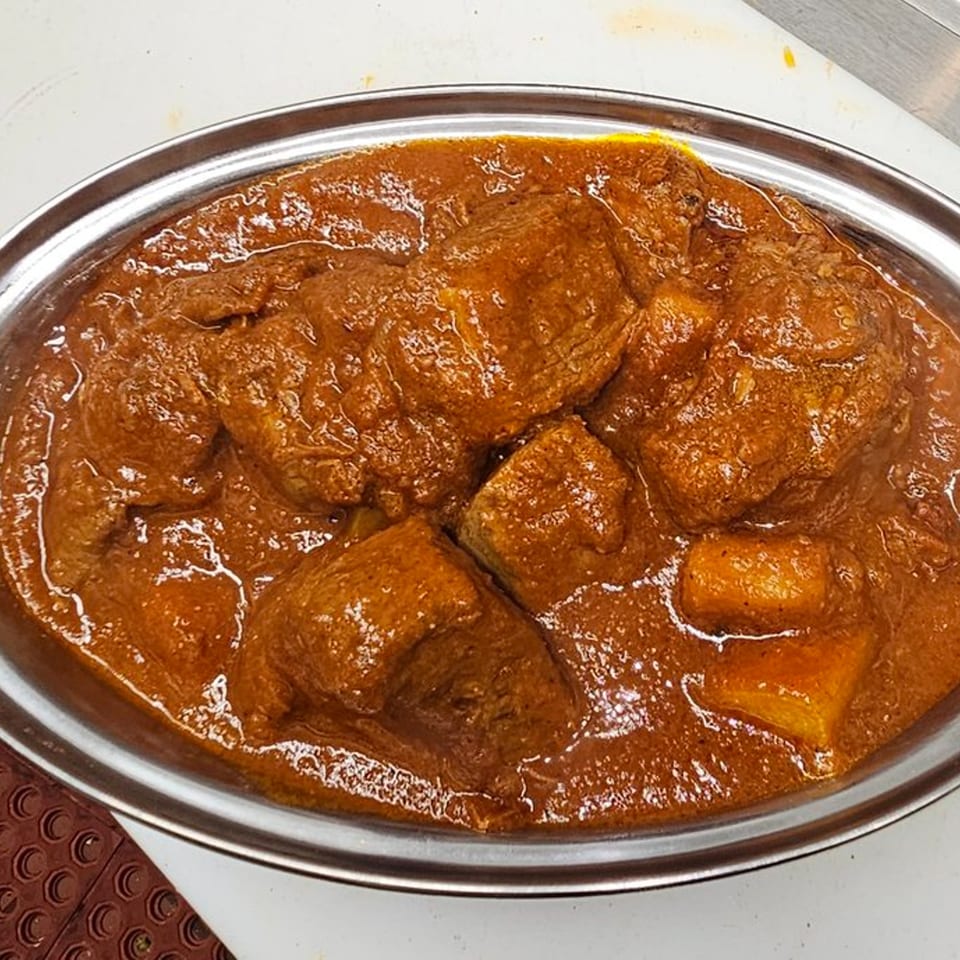 Close-up of a silver bowl filled with rich, reddish-brown Indian curry and large pieces of meat.